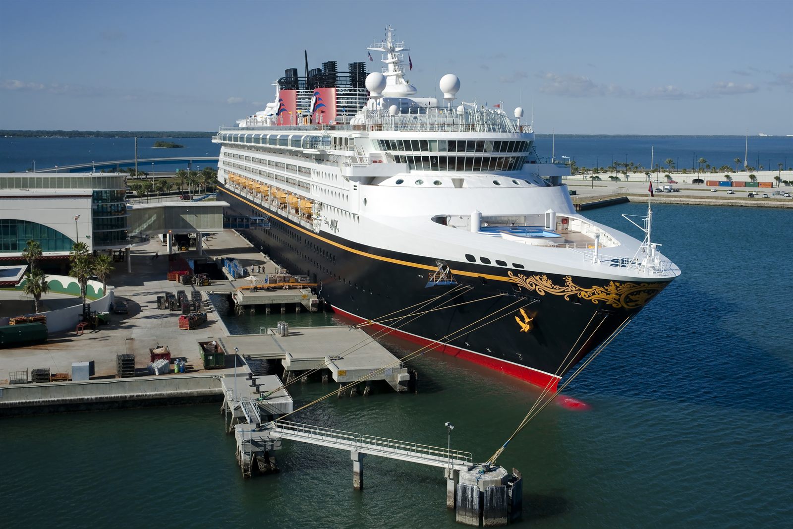 A Disney cruise ship docked beside a cruise terminal on a sunny day; to find a travel agent for cruises, look for one who knows the line