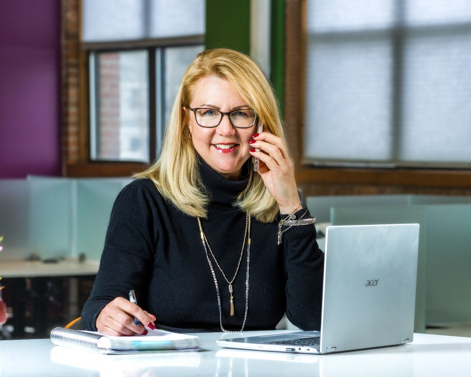 A smiling woman with glasses on the phone at a desk with a laptop and notebook