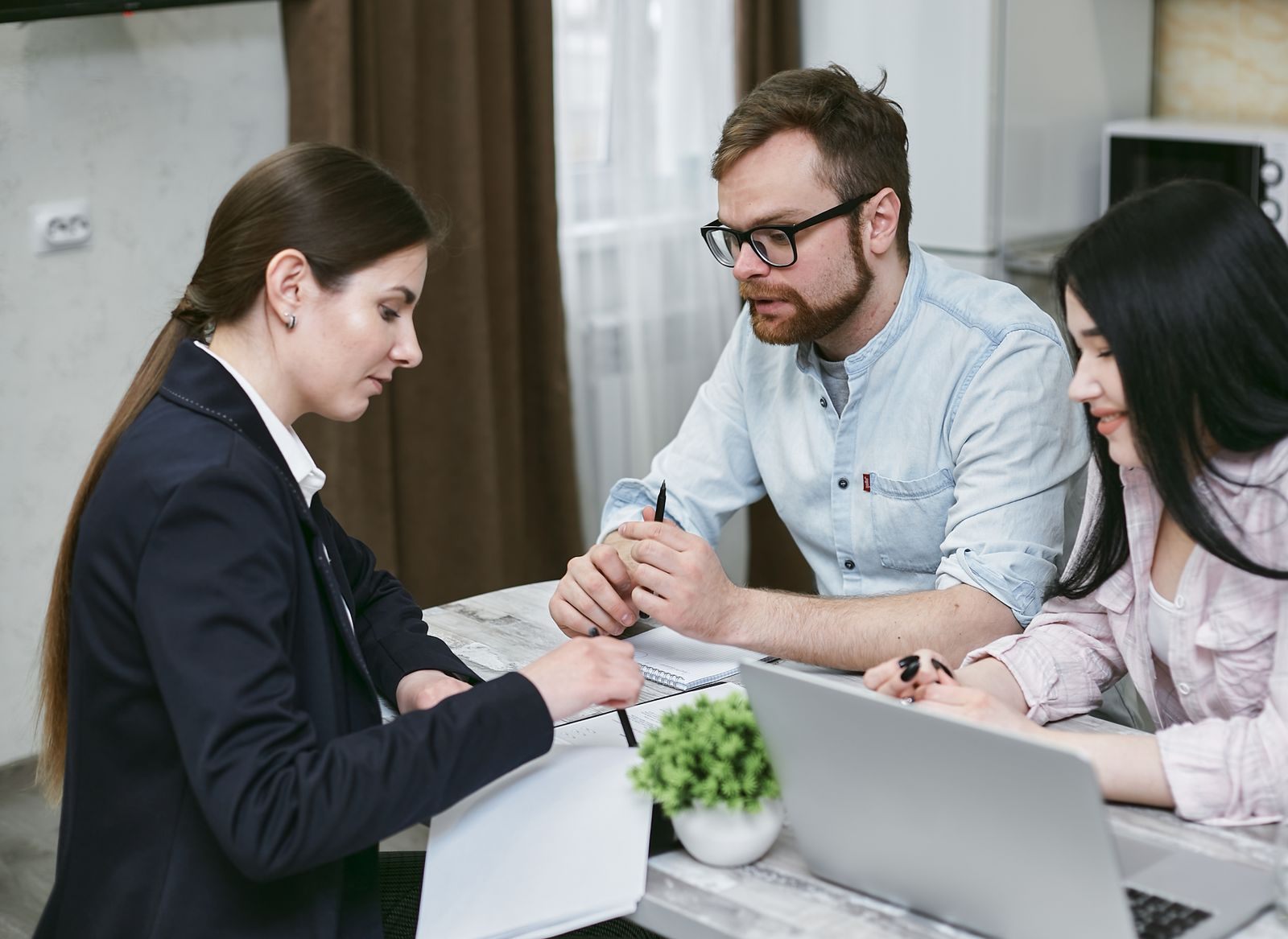 An advisor in a dark blazer going over papers with a couple at a desk with a laptop, the kind of meeting that shows how to choose a travel agent