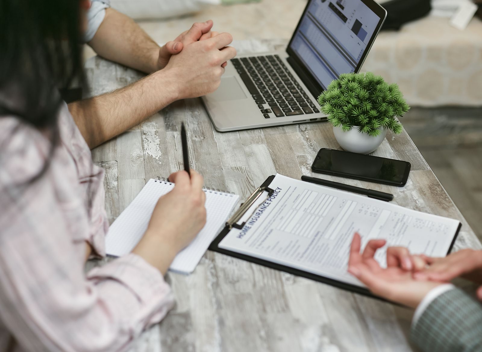 Hands over a clipboard form, a notebook and a laptop on a wooden table during a meeting